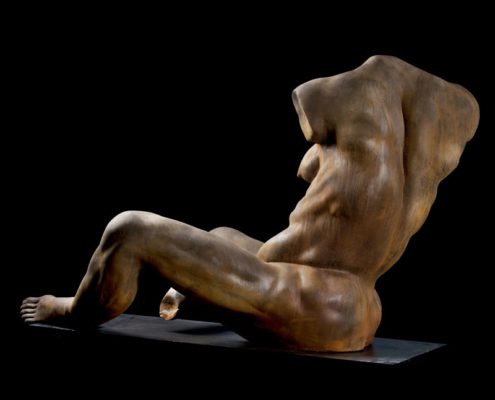 A naked man sitting on the ground in front of a black background.